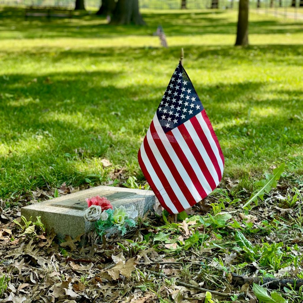 A solemn scene with an American flag beside a tombstone in a grassy cemetery.